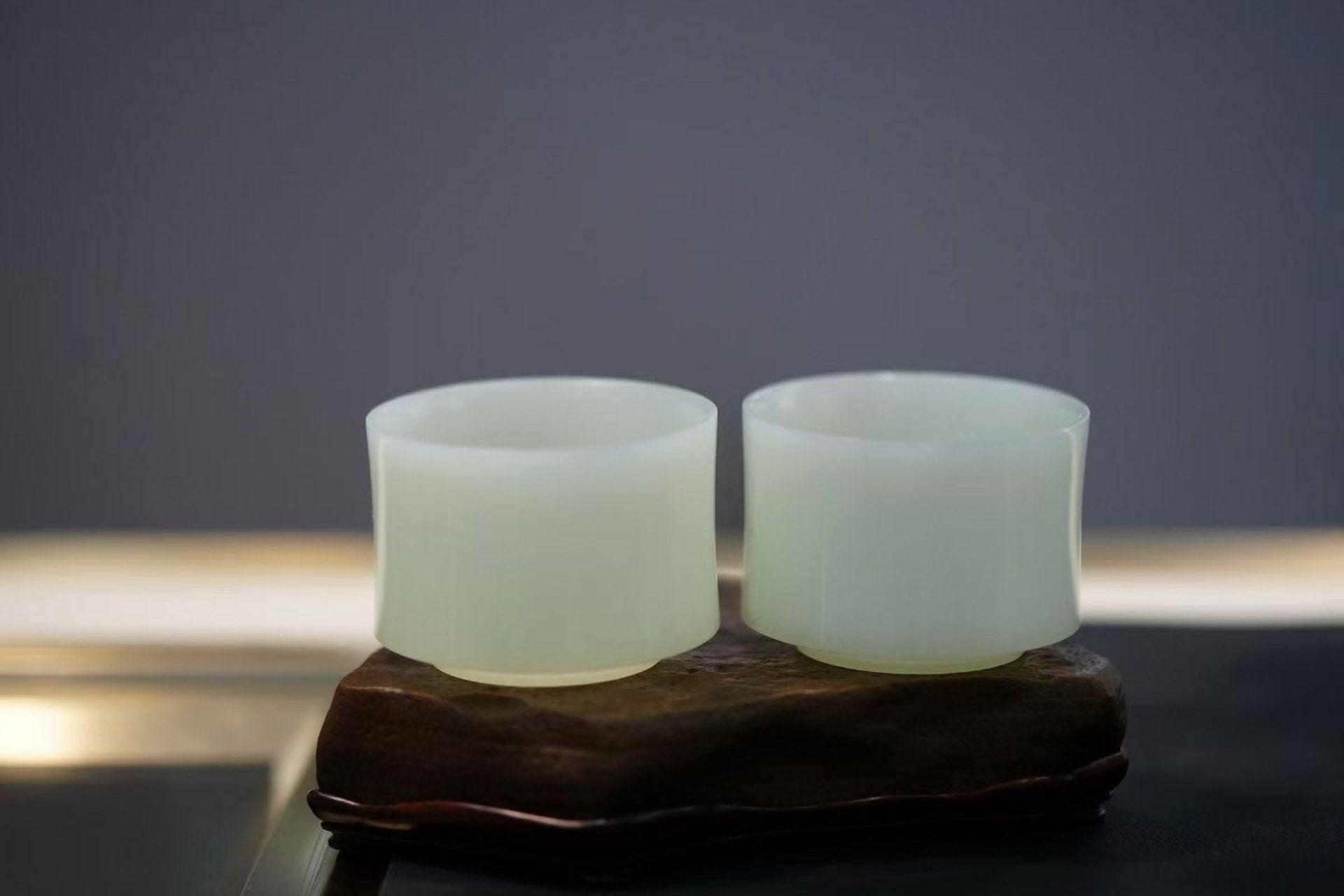 Two pale green-white cups with gently waisted sides, set on a dark wooden stand against a grey wall