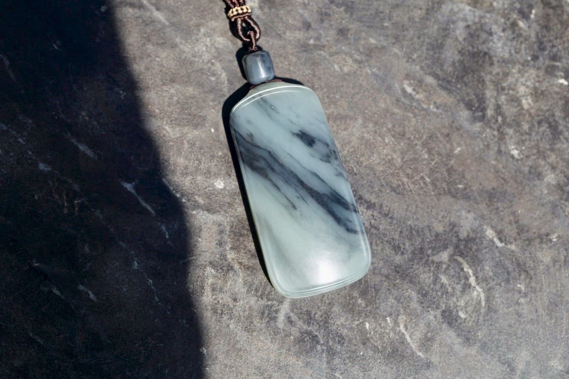 Pale grey-green rectangular pendant with dark ink-like streaks and a barrel bead above, on a sunlit grey surface