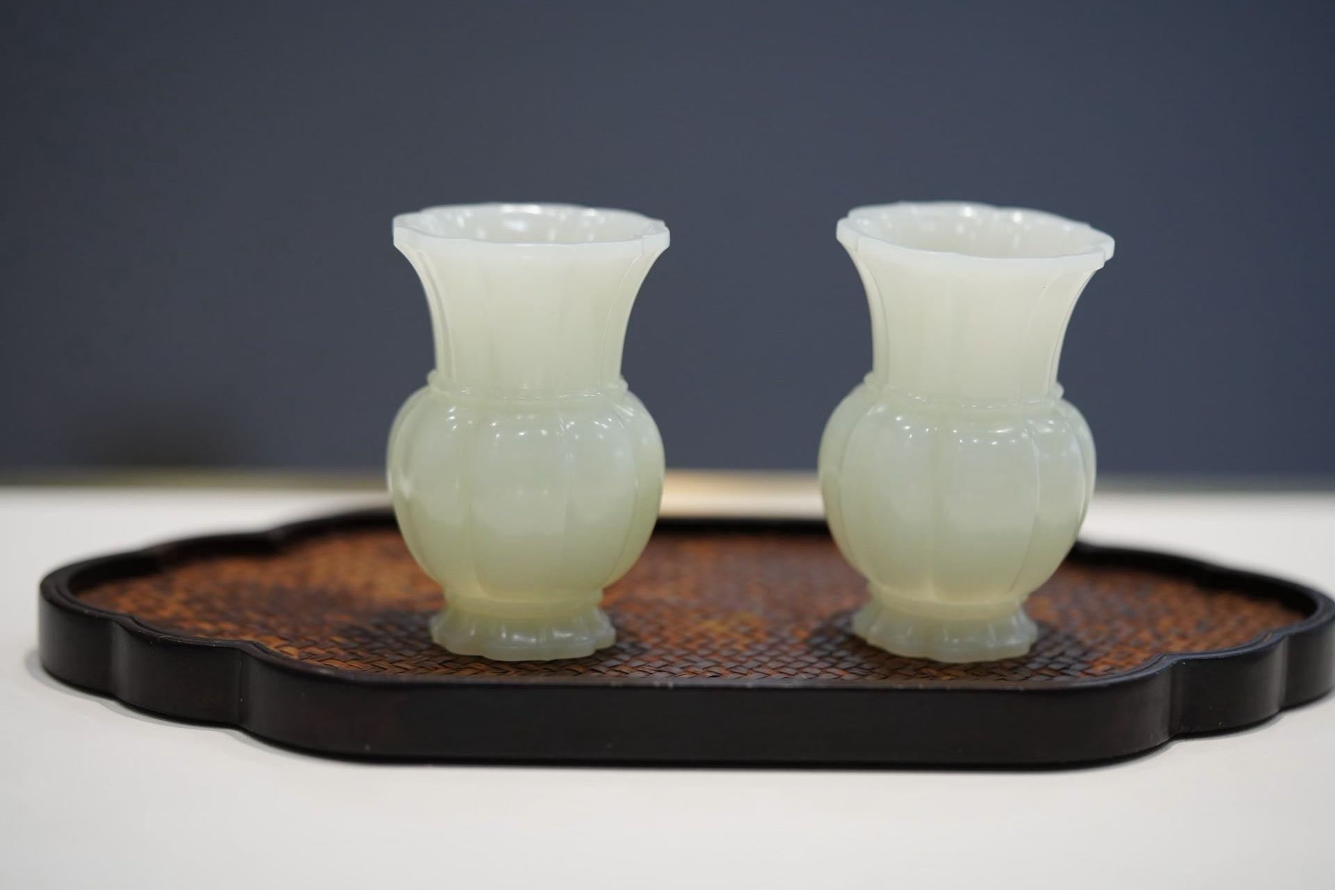 A pair of small pale green-white vases with flared rims and lobed bodies, on a dark tray with a woven centre