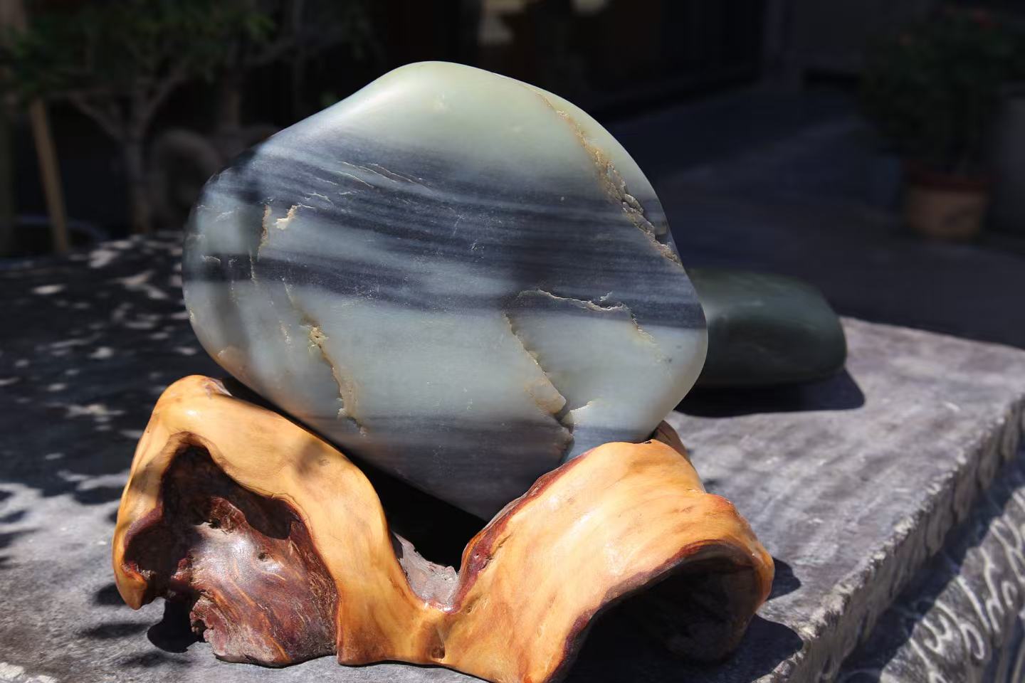 A large stone banded pale green and dark grey, set on a root-wood stand outdoors in sunlight, a dark green stone behind